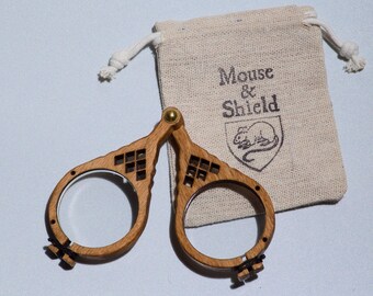 May include: A pair of folding wooden magnifying glasses with circular lenses and a brass hinge, accompanied by a small beige drawstring pouch featuring the printed text Mouse & Shield above a line drawing of a mouse inside a shield emblem.