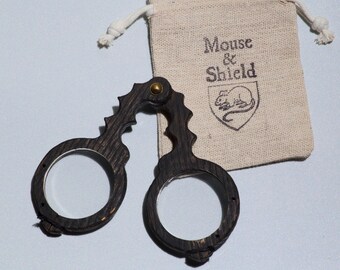 May include: A dark brown wooden handheld magnifying glass with two circular lenses and a serrated handle, joined by a brass rivet. It is accompanied by a beige drawstring fabric pouch featuring the printed text Mouse & Shield and a mouse icon.