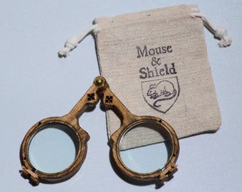 May include: A pair of folding spectacles with circular wooden frames and clear lenses, connected by a central brass hinge. Beside the glasses is a small, beige drawstring pouch made of fabric, featuring the text Mouse & Shield and a mouse icon.