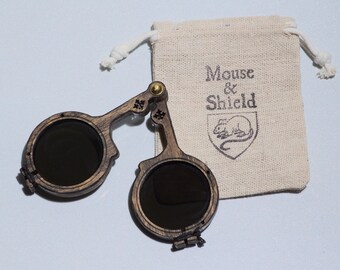 May include: A pair of folding circular sunglasses with dark lenses and a dark brown wood-grain frame, featuring a brass hinge mechanism. Beside the glasses is a small beige fabric drawstring pouch with the text Mouse & Shield and a mouse icon.