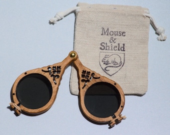 May include: A pair of folding wooden spectacles with dark circular lenses and intricate cut-out details on the frames, connected by a brass hinge. Beside the glasses is a small beige drawstring pouch featuring the text Mouse & Shield.
