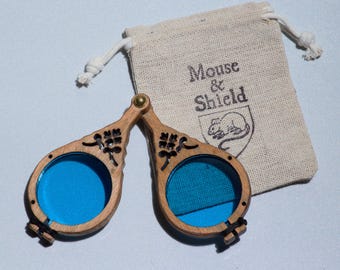 May include: A pair of folding wooden spectacles with circular frames and bright blue lenses, featuring intricate cut-out floral details at the hinges. The glasses are accompanied by a beige drawstring pouch printed with the text Mouse & Shield.