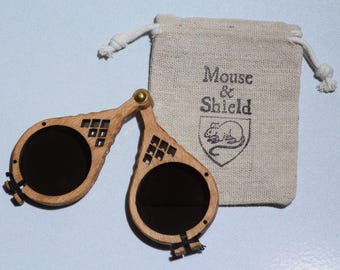 May include: A pair of folding spectacles with circular dark lenses and a light brown wooden frame featuring geometric cutouts. The glasses are accompanied by a small beige drawstring pouch printed with the text Mouse & Shield and a mouse icon.