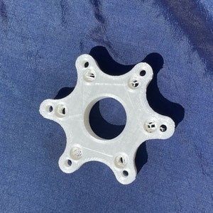 Puede incluir: Un objeto de plástico blanco en forma de estrella de 6 puntas con un agujero en el centro. El objeto tiene una superficie texturizada y parece estar impreso en 3D.