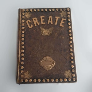 Könnte beinhalten: Ein braunes Lederjournal mit goldenen Akzenten, darunter ein Schmetterling, ein dekoratives Etikett und das Wort "CREATE" in goldenen Buchstaben. Das Journal ist mit goldenen Nieten an den Rändern verziert.
