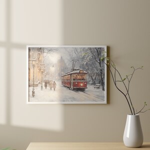 May include: Framed art print of a winter scene featuring a red trolley car on a snow-covered street. The artwork depicts figures walking along the sidewalk, with buildings and trees in the background. The painting has a soft, muted color palette.
