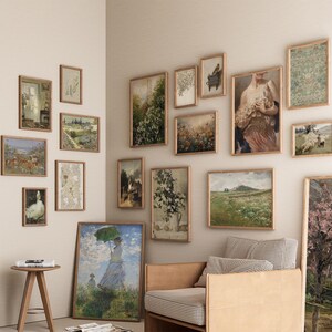 Op de afbeelding: Een galeriewand met een verscheidenheid aan ingelijste kunstwerken, waaronder landschappen, bloemenprints en een portret. De muur is geverfd in een lichtbeige kleur en de lijsten zijn allemaal in een vergelijkbare stijl. Op de voorgrond staat een bruine leren fauteuil met een geruit stoffen kussen.