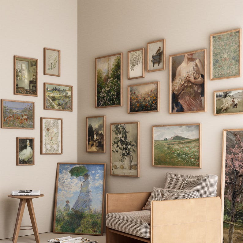 Op de afbeelding: Een galeriewand met een verscheidenheid aan ingelijste kunstwerken, waaronder landschappen, bloemenprints en een portret. De muur is geverfd in een lichtbeige kleur en de lijsten zijn allemaal in een vergelijkbare stijl. Op de voorgrond staat een bruine leren fauteuil met een geruit stoffen kussen.