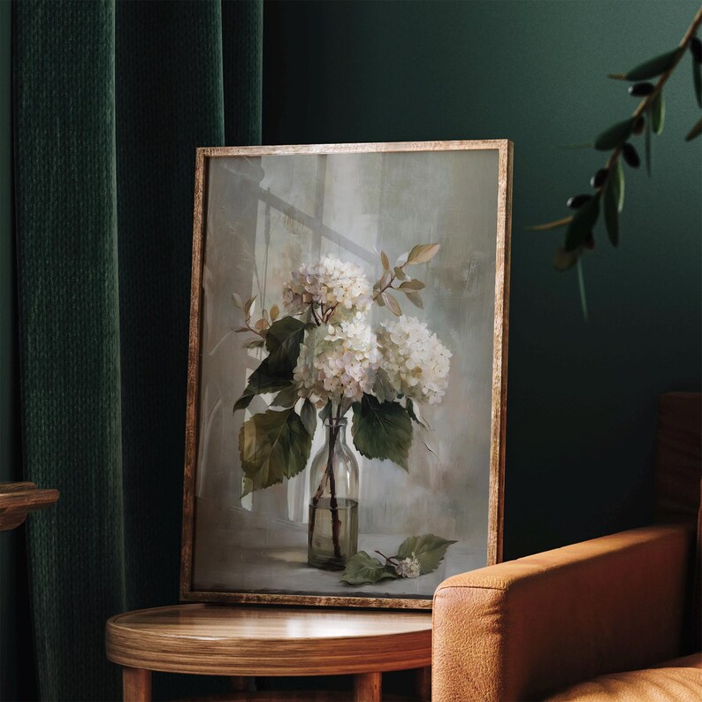 May include: A framed floral print featuring white hydrangeas in a clear glass bottle. The artwork has a gold-toned frame and is set against a muted, neutral background. Green leaves and stems add to the natural aesthetic.