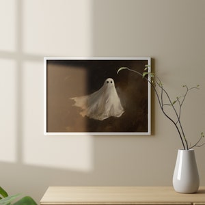 Puede incluir: Una impresi&oacute;n enmarcada con una ilustraci&oacute;n de un fantasma. El fantasma es blanco con dos ojos negros, sobre un fondo marr&oacute;n oscuro. La obra de arte est&aacute; en un marco blanco, contrastando con la pared de tonos c&aacute;lidos.