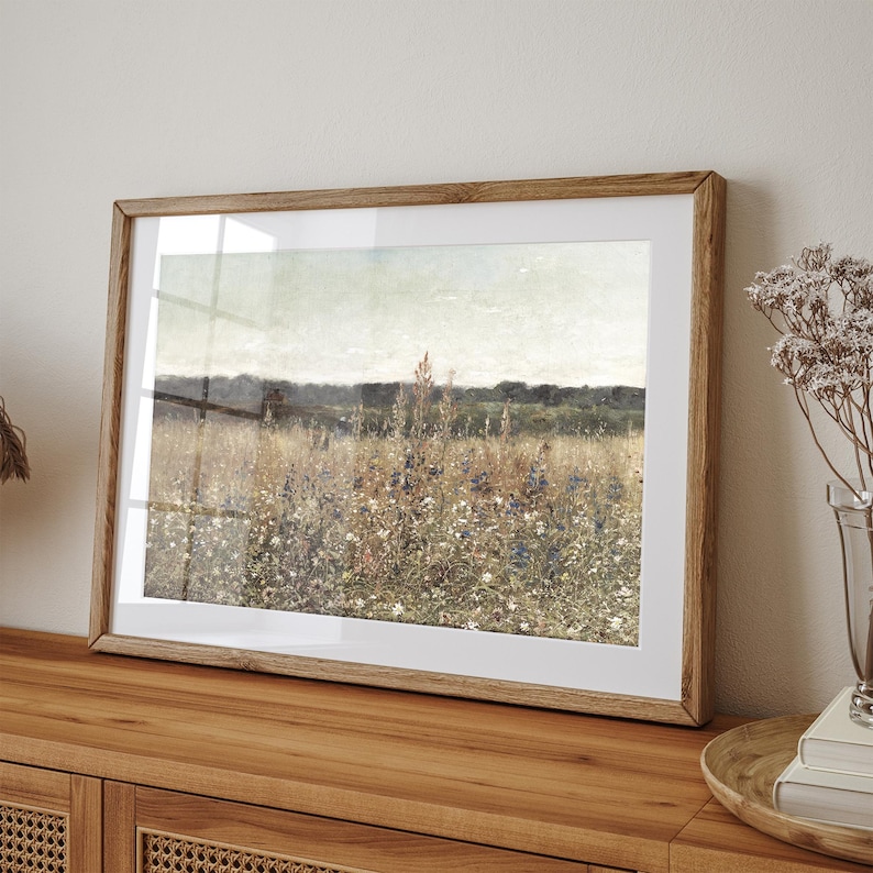 May include: A framed print of a landscape painting depicting a field of wildflowers in shades of blue, green, and white. The painting is set against a backdrop of a distant forest line and a cloudy sky.