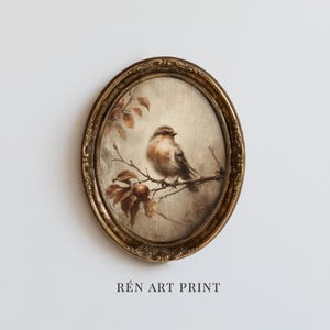 May include: An oval art print featuring a small brown bird perched on a thin branch with leaves and a single round fruit. The artwork is encased in an ornate, textured gold-toned frame. The text REN ART PRINT is displayed below the frame.