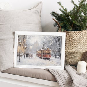 May include: A framed winter landscape painting featuring a red trolley car on a snow-covered street. The artwork, set in a white frame, depicts figures walking along the sidewalk, with buildings and trees in the background. A cozy winter scene.