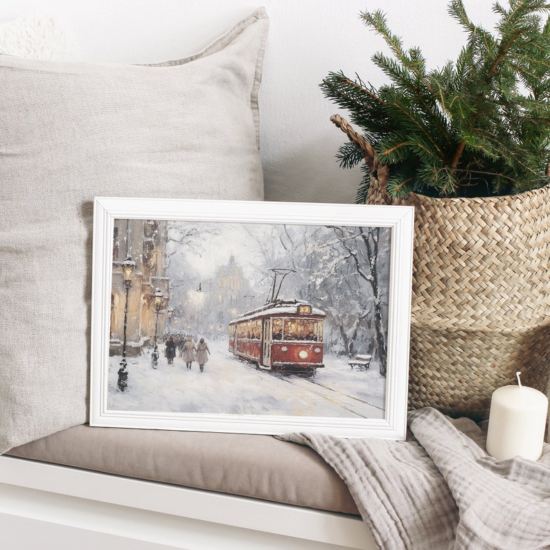 May include: A framed winter landscape painting featuring a red trolley car on a snow-covered street. The artwork, set in a white frame, depicts figures walking along the sidewalk, with buildings and trees in the background. A cozy winter scene.