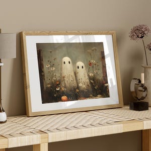 May include: Framed art print featuring two whimsical ghost figures in a field of wildflowers and a small pumpkin. The artwork is set against a muted background, framed in light wood, and evokes a Halloween theme.
