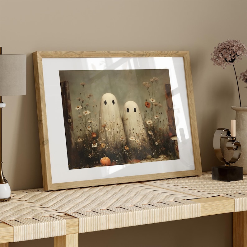 May include: Framed art print featuring two whimsical ghost figures in a field of wildflowers and a small pumpkin. The artwork is set against a muted background, framed in light wood, and evokes a Halloween theme.