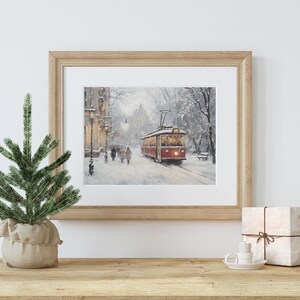 May include: A framed print of a winter scene featuring a red trolley car on a snow-covered street. The artwork is in a light wooden frame and is displayed on a wooden surface with a small Christmas tree and wrapped gift.