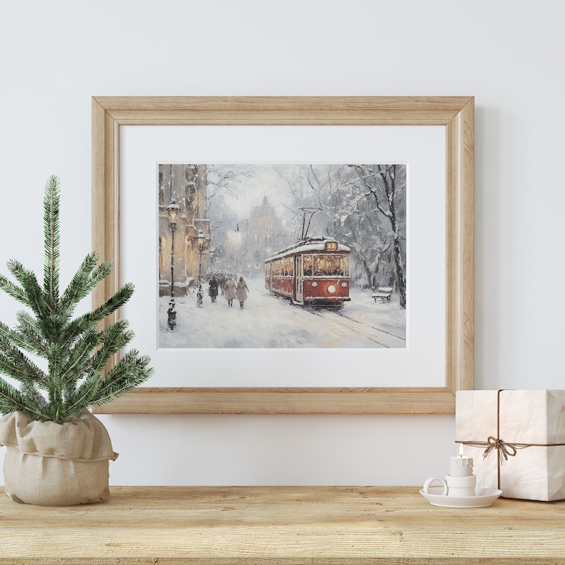 May include: A framed print of a winter scene featuring a red trolley car on a snow-covered street. The artwork is in a light wooden frame and is displayed on a wooden surface with a small Christmas tree and wrapped gift.