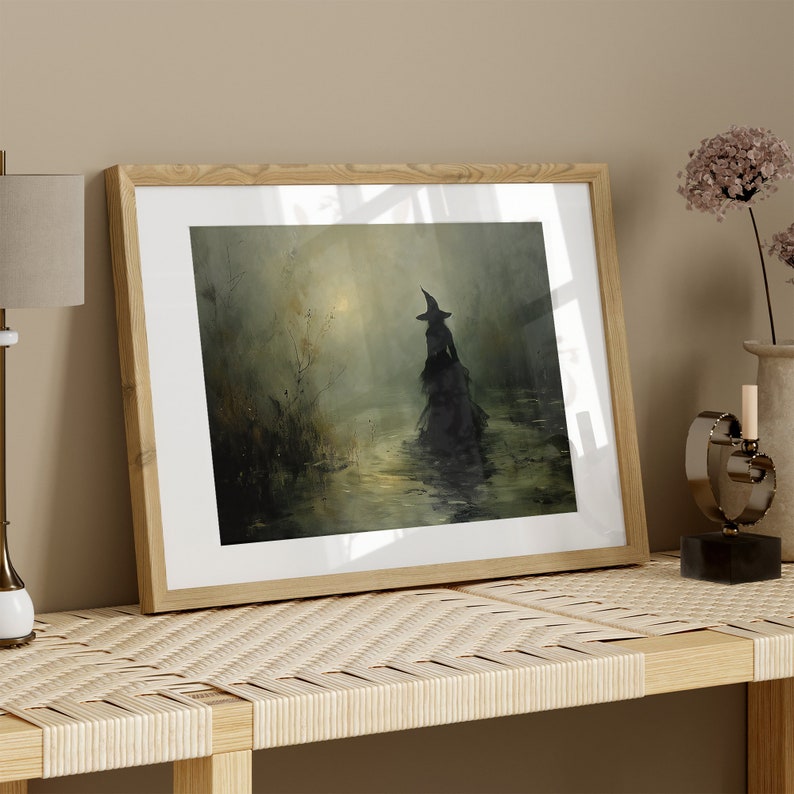 May include: Framed art print featuring a silhouetted figure in a witch's hat and flowing gown, wading through water. The artwork has a dark, atmospheric quality with muted greens and yellows, set in a wooden frame.