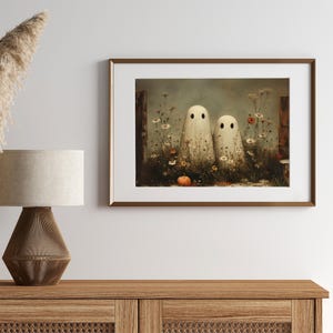 May include: A framed art print featuring two white ghost figures with black eyes standing in a field of wildflowers and a small orange pumpkin. The artwork has a vintage, slightly muted color palette and is set against a soft, atmospheric background.