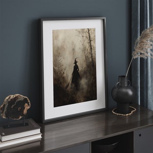 May include: Framed art print depicting a witch's silhouette in a misty forest, rendered in brown and grey tones. The artwork is set within a black frame. The print is displayed on a dark wood surface, alongside decorative items.