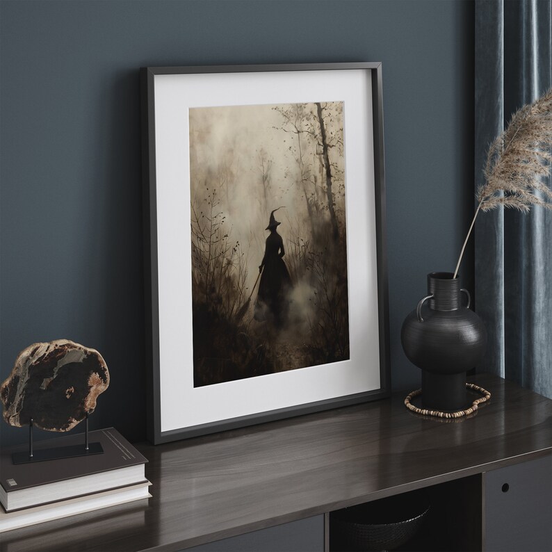 May include: Framed art print depicting a witch's silhouette in a misty forest, rendered in brown and grey tones. The artwork is set within a black frame. The print is displayed on a dark wood surface, alongside decorative items.