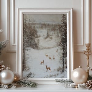 Op de afbeelding: Een ingelijste winterlandschapsschilderij met een besneeuwde bossc&egrave;ne met verschillende herten. Het kunstwerk staat in een witte lijst en is versierd met kerstversieringen en dennentakken.