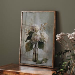 May include: A framed print of white hydrangeas in a clear glass bottle. The artwork features a soft, muted color palette with green leaves and a light background. The frame is a light brown color.
