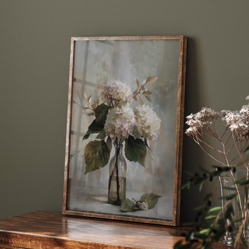 May include: A framed print of white hydrangeas in a clear glass bottle. The artwork features a soft, muted color palette with green leaves and a light background. The frame is a light brown color.