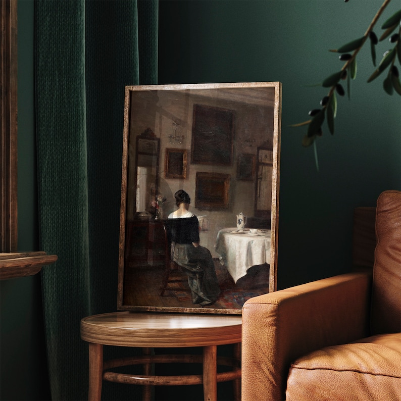 May include: A framed painting depicting a woman in a dark dress sitting at a table in a room with several other paintings on the walls. The painting is in a wooden frame and is hanging on a wall.