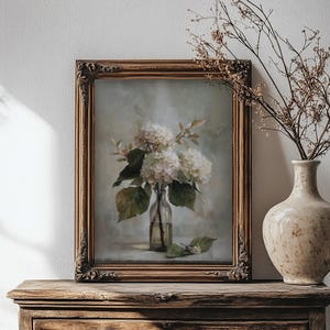 May include: A framed floral still life painting featuring white hydrangeas in a clear glass bottle. The painting has a gold ornate frame and is displayed on a wooden surface. A beige vase with dried branches is to the right.