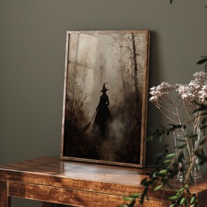 May include: A framed art print showing a silhouetted witch in a misty forest. The witch is wearing a pointed hat and carrying a broom. The artwork uses brown and grey tones, with a vintage style. The frame is made of wood.