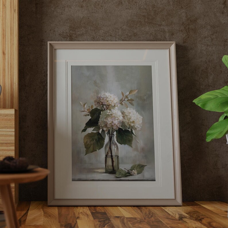 May include: Framed artwork featuring a still life of white hydrangea flowers in a clear glass bottle. The painting has a soft, muted color palette with green leaves and a neutral background. The frame is a light beige color.