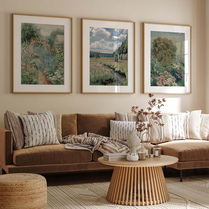 Op de afbeelding: Drie ingelijste landschapschilderijen hangen aan een beige muur boven een bruine leren bank met kussens en een deken. Een houten salontafel staat voor de bank.