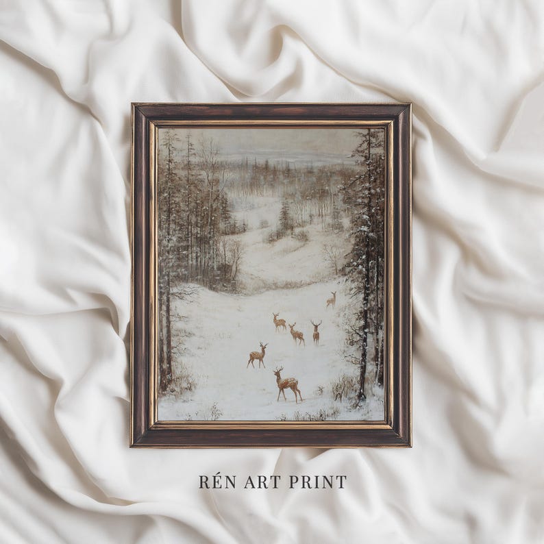 Op de afbeelding: Een ingelijste winterlandschap print met herten in een besneeuwd bos. Het kunstwerk heeft een donkerbruine lijst en de tekst "R&Eacute;N ART PRINT" onderaan. Het kleurenpalet is gedempt met tinten bruin, wit en grijs.