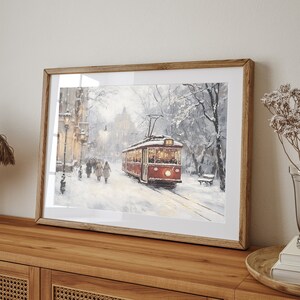 May include: Framed art print depicting a snowy winter scene with a red trolley car on tracks. The artwork features a street lined with buildings and people walking in the snow. The print is encased in a wooden frame and displayed on a wooden cabinet.