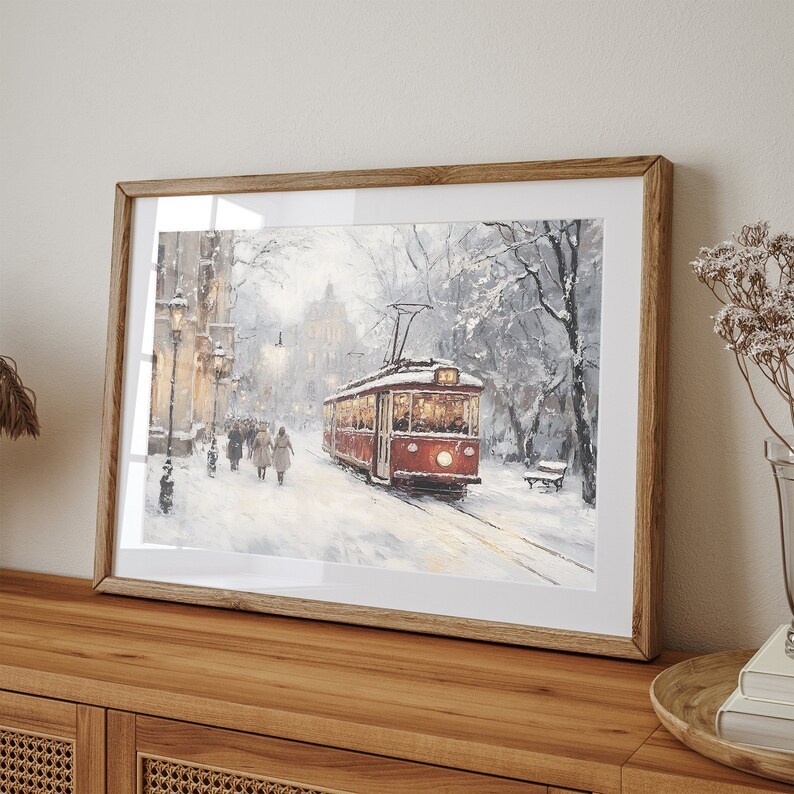 May include: Framed art print depicting a snowy winter scene with a red trolley car on tracks. The artwork features a street lined with buildings and people walking in the snow. The print is encased in a wooden frame and displayed on a wooden cabinet.