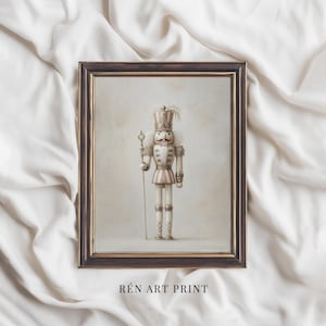 May include: A framed print of a vintage nutcracker soldier. The artwork shows a detailed, realistic nutcracker in a white and pink striped uniform, a tall hat, and a mustache. The frame is dark brown. The text "RÉN ART PRINT" is at the bottom.