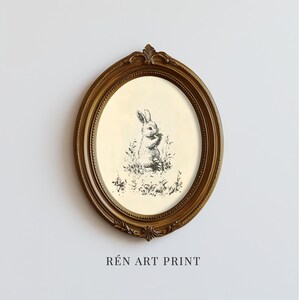 May include: An oval framed art print featuring a black and white illustration of a rabbit in a field of grass and flowers. The frame is a dark gold color with ornate detailing. The text "R&Eacute;N ART PRINT" is printed at the bottom.
