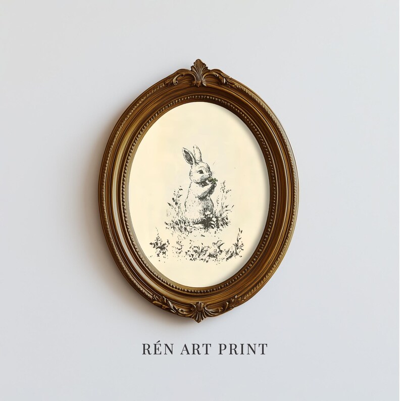 May include: An oval framed art print featuring a black and white illustration of a rabbit in a field of grass and flowers. The frame is a dark gold color with ornate detailing. The text "R&Eacute;N ART PRINT" is printed at the bottom.