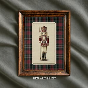 May include: A framed art print featuring a nutcracker soldier in a red and gold uniform holding a spear. The nutcracker is centered on a cream-colored background, which is layered over a dark green and red plaid pattern. The frame is metallic bronze.