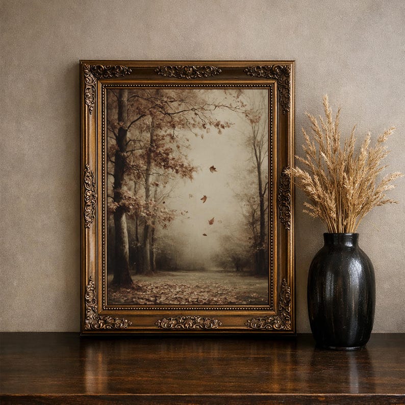 May include: A rectangular wall art piece in an ornate, antique bronze-colored frame featuring a sepia-toned forest scene with falling autumn leaves. Beside the frame sits a black ceramic vase filled with dried, light brown ornamental grasses.