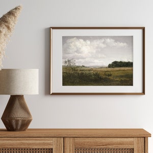 Pode incluir: Uma impress&atilde;o de arte emoldurada com uma cena pastoral com um c&eacute;u nublado. A obra de arte retrata um campo com &aacute;rvores e vegeta&ccedil;&atilde;o, emoldurada em uma moldura de metal dourado. A impress&atilde;o &eacute; exibida em um arm&aacute;rio de madeira, ao lado de uma l&acirc;mpada com uma abajur bege.