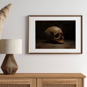 May include: A framed painting of a human skull in a dark setting. The skull is positioned on a surface and is facing to the right. The skull is a light brown color and has a detailed texture.
