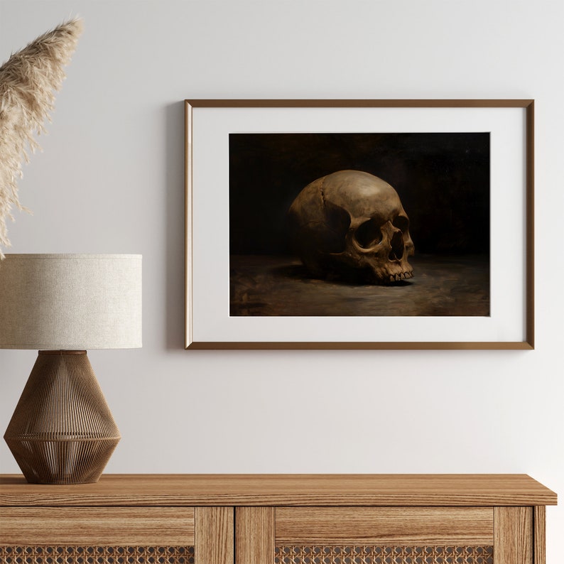 May include: A framed painting of a human skull in a dark setting. The skull is positioned on a surface and is facing to the right. The skull is a light brown color and has a detailed texture.