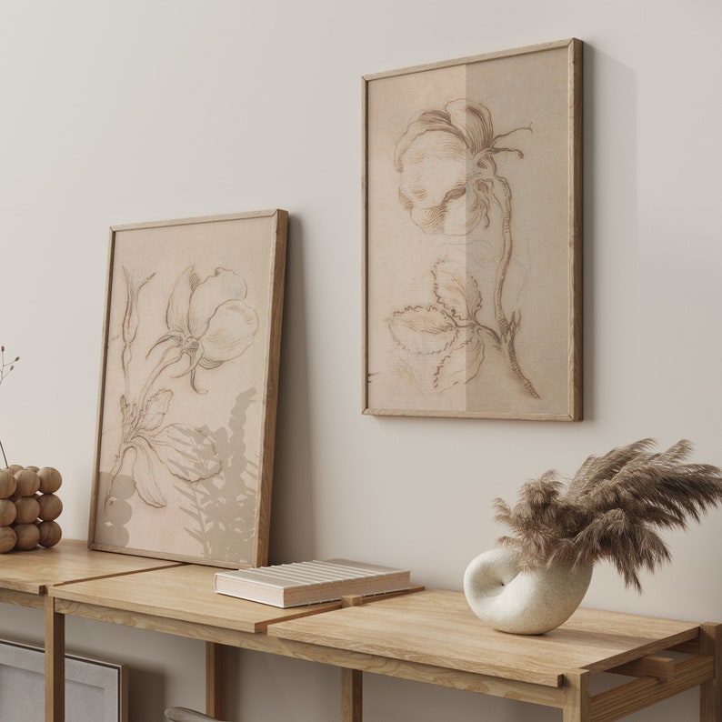 May include: Two framed botanical illustrations of flowers in shades of brown on a white wall. The frames are simple and wooden. A wooden shelf with a book and a white ceramic vase with dried pampas grass sits below the frames.