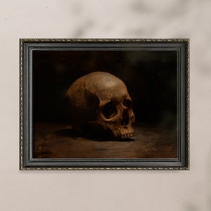 May include: A framed painting of a human skull in a dark brown setting. The skull is positioned on a dark brown surface and is facing to the right. The painting is framed in a black frame with a gold trim.