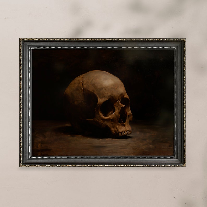 May include: A framed painting of a human skull in a dark brown setting. The skull is positioned on a dark brown surface and is facing to the right. The painting is framed in a black frame with a gold trim.
