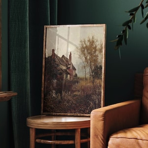 May include: A framed wall art print featuring a landscape painting of a rustic cottage with a chimney, surrounded by tall trees and overgrown vegetation in muted earth tones of brown, beige, and dark green, set within a thin metallic frame.