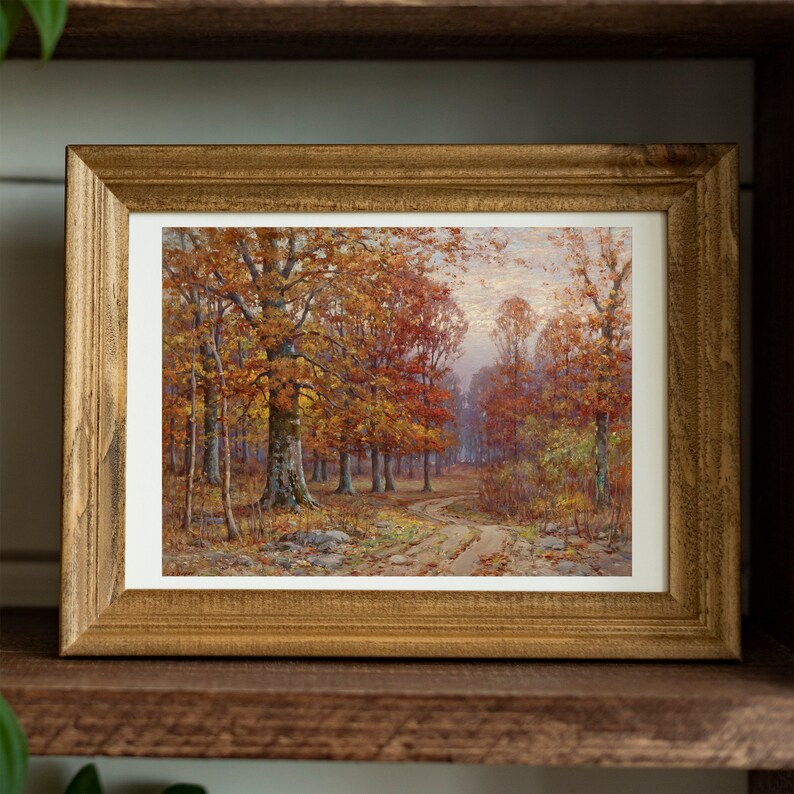May include: A framed landscape painting of an autumn forest scene. The artwork features a dirt path winding through trees with vibrant orange and red leaves. The frame is made of rustic, light brown wood.
